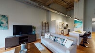 Located at 180 E Main St in Haw River, NC, this single-floor space showcases a compelling blend of historic industrial character and contemporary design. Soaring ceilings with exposed reclaimed wood beams and visible ductwork establish an authentic loft aesthetic throughout, while warm hardwood flooring grounds each area cohesively.

The open living area is anchored by a striking exposed brick wall with arched windows that flood the space with natural light. The generous layout accommodates distinct seating zones, with ceiling fans adding to the airy feel. The kitchen features white shaker cabinetry, stainless steel appliances, and a sleek quartz-topped island with bar seating, illuminated by industrial-style pendant lights that complement the overall design language.

A hallway leads to the bedroom, which maintains the same lofty ceiling height with exposed beams overhead and hardwood floors underfoot, offering a calm, restful atmosphere. A welcoming entryway nook with a console table provides a refined transition into the space.

Throughout, the interplay of raw architectural elements — weathered brick, aged timber, and exposed mechanicals — paired with polished modern finishes creates a dynamic, character-rich environment.