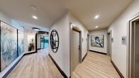 Located at Reserve Trail in Jensen Beach, Florida, this single-story fitness facility is thoughtfully designed to deliver a premium workout experience. The main gym area is defined by soaring ceilings with exposed wood beam detailing and expansive floor-to-ceiling windows that flood the space with natural light and frame views of the surrounding tropical landscape. The open layout accommodates a comprehensive selection of cardio equipment — including treadmills, ellipticals, stationary bikes, and rowing machines — as well as an extensive strength training section featuring free weights, kettlebells, and cable systems. Bold geometric patterned wall art and large mirrored panels add visual depth and a modern aesthetic throughout. A separate studio room features a distinctive metallic brick accent wall, a skylight, a statement chandelier, and ample open floor space equipped with stability balls and resistance equipment. The connecting corridor is finished with light wood-look flooring, recessed lighting, a decorative round mirror, and nature-inspired artwork, giving the facility a polished, welcoming feel from entry to exit.