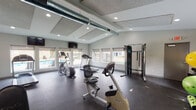Located in Portage, Michigan, this single-floor fitness center is designed to support a comprehensive workout experience. The main exercise area is generously sized, featuring durable rubber flooring that accommodates a wide range of equipment, including multiple treadmills, an elliptical trainer, a stationary bike, and a cable/functional trainer machine. A dedicated section hosts a dumbbell rack alongside stability balls, offering space for free weight and core training. Large windows line the walls, flooding the room with natural light and providing views of the adjacent outdoor pool area, creating an energizing atmosphere during workouts. Wall-mounted flat-screen TVs are positioned throughout for entertainment. Exposed industrial-style ductwork runs along the ceiling, complementing the clean, modern aesthetic of the neutral-toned walls and recessed lighting. Full-length mirrors expand the sense of space and allow for form monitoring during exercise. A separate transitional area includes built-in open lockers with bench seating, a water fountain, and access to restroom facilities, providing practical amenities for users. The space is bright, functional, and thoughtfully laid out to serve a variety of fitness needs.