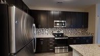 This single-floor space showcases a thoughtfully designed interior with a strong emphasis on modern aesthetics and functional living. The kitchen stands out as a centerpiece, outfitted with rich, dark espresso cabinetry that extends from floor to ceiling, maximizing storage while creating a bold, sophisticated look. Stainless steel appliances — including a full-size refrigerator, a range with oven, and an over-the-range microwave — lend a polished, contemporary finish throughout the space. Light granite countertops provide a striking contrast against the dark cabinetry, offering ample workspace for everyday use. A mosaic tile backsplash featuring a blend of neutral tones and subtle metallic accents adds a refined textural detail that ties the design together seamlessly. The cabinetry is accented with sleek bar-pull hardware in a brushed chrome finish, reinforcing the modern style. Wood-look flooring grounds the space with warmth while complementing the overall color palette. The layout is efficient and well-organized, reflecting a design approach that balances form and function. Every detail, from the material choices to the hardware finishes, contributes to a cohesive, upscale atmosphere throughout this inviting space.