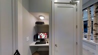 This single-floor business center is designed to provide a focused and functional workspace in a compact, efficiently organized layout. The space centers around a built-in desk nook tucked neatly into a recessed alcove, making smart use of the available area. The workstation is equipped with a desktop computer, a full-sized monitor, keyboard, and mouse, offering the essentials for productivity. An upper shelf positioned above the desk houses a printer, keeping equipment organized and within easy reach without cluttering the work surface.

The interior features neutral gray walls that contribute to a calm, distraction-free environment suited for focused tasks. A flush-mounted ceiling light provides direct, functional illumination over the workspace. The space is accessed through a clean, white panel door fitted with a standard lever handle, reinforcing the straightforward, utilitarian design of the area. The overall aesthetic is minimal and purposeful, prioritizing practicality over ornamentation. This business center serves as a convenient, self-contained resource for those needing quick access to computing and printing facilities.
