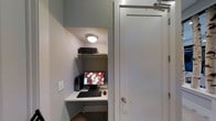 This single-floor business center is designed to provide a focused and functional workspace in a compact, efficiently organized layout. The space centers around a built-in desk nook tucked neatly into a recessed alcove, making smart use of the available area. The workstation is equipped with a desktop computer, a full-sized monitor, keyboard, and mouse, offering the essentials for productivity. An upper shelf positioned above the desk houses a printer, keeping equipment organized and within easy reach without cluttering the work surface.

The interior features neutral gray walls that contribute to a calm, distraction-free environment suited for focused tasks. A flush-mounted ceiling light provides direct, functional illumination over the workspace. The space is accessed through a clean, white panel door fitted with a standard lever handle, reinforcing the straightforward, utilitarian design of the area. The overall aesthetic is minimal and purposeful, prioritizing practicality over ornamentation. This business center serves as a convenient, self-contained resource for those needing quick access to computing and printing facilities.