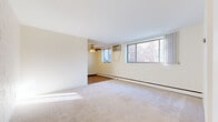 This single-floor unit offers a practical and comfortable layout across several well-defined living spaces. The main living and dining area is generously proportioned, featuring neutral wall-to-wall carpeting, bright white walls, and large windows that invite ample natural light while offering views of the surrounding brick exterior. A ceiling fan provides additional comfort, and a baseboard heating system runs along the perimeter walls. An adjacent secondary room, also carpeted and ceiling fan-equipped, offers versatility as a bedroom or flex space with its own window and natural light. The bedroom area includes built-in shelving for added storage alongside closet space, with warm wood-trimmed doors throughout adding a classic touch. The galley-style kitchen is well-equipped, featuring white cabinetry, granite-patterned countertops, a double sink, and white appliances including a gas range and full-size refrigerator. Rich hardwood flooring in the kitchen adds warmth and contrast. The bathroom is finished in clean, white square tiles from floor to ceiling, with a full bathtub and shower, a classic wall-mounted sink, basket-weave tile flooring, and a mirrored medicine cabinet above, completing this straightforward and functional residential space.