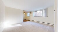 This single-floor unit offers a practical and comfortable layout across several well-defined living spaces. The main living and dining area is generously proportioned, featuring neutral wall-to-wall carpeting, bright white walls, and large windows that invite ample natural light while offering views of the surrounding brick exterior. A ceiling fan provides additional comfort, and a baseboard heating system runs along the perimeter walls. An adjacent secondary room, also carpeted and ceiling fan-equipped, offers versatility as a bedroom or flex space with its own window and natural light. The bedroom area includes built-in shelving for added storage alongside closet space, with warm wood-trimmed doors throughout adding a classic touch. The galley-style kitchen is well-equipped, featuring white cabinetry, granite-patterned countertops, a double sink, and white appliances including a gas range and full-size refrigerator. Rich hardwood flooring in the kitchen adds warmth and contrast. The bathroom is finished in clean, white square tiles from floor to ceiling, with a full bathtub and shower, a classic wall-mounted sink, basket-weave tile flooring, and a mirrored medicine cabinet above, completing this straightforward and functional residential space.