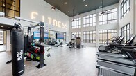 Located at 4555 McCrimmon Pkwy in Morrisville, NC, this fitness center is a standout facility defined by its dramatic double-height ceilings, abundant natural light, and a clean, contemporary aesthetic. The main workout area greets visitors with an illuminated "FITNESS" neon sign and a bright, open floor plan finished with light wood-look flooring and crisp white walls accented by a dark ceiling detail.

Floor-to-ceiling black-framed windows wrap the space, offering views of the surrounding outdoor amenity areas and creating an airy, energizing atmosphere. The main floor houses a robust selection of strength and functional training equipment, including a multi-station rig with TRX suspension trainers, a heavy bag, kettlebells, rowing machines, and selectorized weight machines. Large wall-mounted displays support on-demand workout programming.

A dedicated studio room, separated by glass-panel sliding doors, provides a quieter space for cycling on spin bikes, balance training with BOSU balls, and functional work with medicine balls and a full rack of dumbbells. Mirrors along the studio wall enhance the sense of space and allow users to monitor their form.

The cardio zone features a row of modern treadmills and stair climbers positioned directly in front of expansive windows, making for an inspiring workout backdrop. Ceiling fans and recessed lighting throughout ensure comfort, while the glass-walled studio and open sightlines keep the entire facility feeling connected and well-organized.