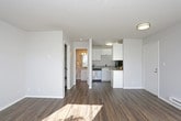 This single-floor unit offers a clean, functional layout designed for efficient everyday living. Throughout the space, rich dark hardwood-style flooring provides a warm contrast against the crisp white walls and trim. A textured ceiling adds visual depth to each room.

The open living area is a standout feature, benefiting from multiple windows that invite abundant natural light, casting bright patterns across the floors. A baseboard heater runs along one wall, and a ceiling-mounted flush light fixture provides additional illumination near the entryway. The front door opens directly into this versatile living space.

The kitchen is positioned centrally within the unit and features white cabinetry offering ample upper and lower storage. Appliances include a black-and-stainless refrigerator, an electric coil range, an over-the-range microwave, and a stainless steel dishwasher. Laminate countertops complete the functional workspace.

Adjacent to the kitchen, a walk-in closet provides generous storage. The full bathroom is neatly appointed with a tub-shower combination, a single-sink vanity with a laminate countertop, and a well-lit mirror above. The overall ambiance of the unit is bright, clean, and move-in ready.