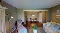 This single-floor residence offers a thoughtfully arranged open-concept layout that seamlessly connects the living and dining areas, creating a welcoming and functional flow throughout the home. The living area is furnished with comfortable seating, including a chaise lounge and sofa, centered around a tiled coffee table accented with lush greenery. Warm, neutral tones in taupe and olive coat the walls, lending a calm and inviting atmosphere to the entire space. Rich, dark hardwood flooring runs through the dining area, contrasting beautifully with the carpeted lounge space and grounding the overall design. The dining room is framed by a partial wall opening, drawing the eye toward a round dining table surrounded by elegant dark wood chairs. Above the table, a classic pendant chandelier adds a refined focal point, while a decorative round mirror on the wall enhances the sense of depth and style. A set of closet doors is visible along the dining room wall, offering practical built-in storage. Natural light filters softly through the space, complementing the warm interior palette. The cohesive design and open sightlines make this a comfortable, well-appointed single-level home ideal for both everyday living and casual entertaining.