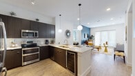 This thoughtfully designed single-floor residence offers a seamless blend of modern aesthetics and functional living. The open-concept kitchen is a standout feature, showcasing rich dark cabinetry, stainless steel appliances including a double oven and French door refrigerator, a white quartz peninsula with an undermount sink, a built-in wine cooler, and globe pendant lighting that adds a contemporary touch. Light wood-look flooring flows throughout, connecting the kitchen to a welcoming dining area with a marble-topped table and through to the living room, which features a bold blue accent wall and large French doors that invite natural light. A separate den or flex room provides additional versatile living space. The primary bedroom is a calming retreat with a soft blue accent wall, ceiling fan, and natural light from large windows. It is complemented by a walk-in closet with built-in shelving and a luxurious en-suite bathroom finished in floor-to-ceiling marble-style tile with a soaking tub. A secondary bathroom mirrors the refined aesthetic with a tub-shower combination and marble-tile surround. Crisp white walls, recessed lighting, and cohesive finishes throughout give the space a polished, contemporary ambiance.