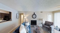 This single-floor space offers a comfortable and well-appointed layout designed for easy living. The living room serves as the heart of the home, centered around a classic fireplace with a clean white surround that adds a touch of warmth and charm. Neutral wall tones and light-colored carpeting create a calm, cohesive backdrop throughout the space. The room is furnished with a plush blue sofa accented by decorative throw pillows, paired with a patterned accent chair, and anchored by a glass-top coffee table — a combination that balances comfort with style. A textured ceiling adds subtle architectural character, while ambient lighting from floor and table lamps creates a soft, welcoming glow. Wall art and a decorative oversized clock contribute to the curated, lived-in feel of the space. A hallway visible from the living area suggests additional rooms beyond, hinting at a practical and functional floor plan. Overall, the space conveys a cozy, approachable aesthetic with classic design elements that make it feel both homey and well-maintained.