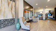 This single-floor community space is designed with a contemporary aesthetic, blending functionality and style across multiple interconnected areas. The main lobby welcomes visitors with light wood-look tile flooring, elegant high-back accent chairs, and decorative elements including a tall teal ceramic vase and geometric lattice wall panels. Frosted glass office partitions with dark frames line one side, creating semi-private meeting spaces while maintaining an open, airy feel.

The lounge area extends into a spacious common room with recessed and pendant lighting, neutral tones, and plush seating arrangements. A wall-mounted flat-screen TV and decorative accents add a polished, residential touch. Transitional flooring shifts between tile and patterned carpet, defining distinct functional zones.

A fully equipped kitchen and dining area features rich dark cabinetry, white quartz countertops, stainless steel appliances, a subway tile backsplash, and a center island with bar-height seating illuminated by pendant lights. Nearby, high-top bistro tables and seating offer an informal gathering spot. Entry vestibules on multiple sides provide natural light through large windows, with views of the surrounding exterior. The overall ambiance is refined, welcoming, and well-appointed throughout.