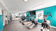Located at 1717 Aberdeen Rd in Towson, Maryland, this dedicated fitness center offers a well-equipped single-room exercise space designed to support a comprehensive workout experience. The room is defined by its vibrant teal accent wall, which contrasts cleanly against the light gray walls and white trim, creating an energetic yet focused atmosphere. Durable interlocking rubber flooring covers the entire space, providing both safety and comfort underfoot during exercise. Natural light streams in through several white-framed windows positioned along the walls, keeping the environment bright and inviting. Overhead, recessed and flush-mounted lighting supplements the natural light for consistent illumination throughout the day. The cardio section is well-stocked with multiple True treadmills and elliptical trainers, all arranged in a row for easy access. A wall-mounted flat-screen television faces the cardio equipment, enhancing the workout experience. The strength training area features a Hoist cable machine, a dumbbell rack with a full range of free weights, and a flat workout bench. Full-length mirrors line one wall, visually expanding the space and assisting with form. Yoga mats, resistance bands, and stability equipment are also available, rounding out the facility's offerings.