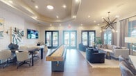 Located in Doral, Florida, this well-appointed community clubroom offers a sophisticated and versatile gathering space spread across a single open floor. The interior is anchored by a distinctive tray ceiling with recessed lighting that adds depth and elegance to the room. Large-format tile flooring runs throughout, providing a clean, modern foundation for the varied activity zones within.

A centerpiece shuffleboard table defines the heart of the space, lending a social and recreational focal point. Flanking it are multiple seating arrangements, including a lounge area with a plush sofa, leather accent chairs, and a tufted navy ottoman, as well as a casual TV lounge with built-in bench seating and a wall-mounted flat-screen. A dedicated workspace or dining area features a conference-style table with swivel chairs, complemented by curated artwork and textured white brick accent walls.

A wet bar with a white countertop and bar stools serves as a convenient refreshment station. A striking sputnik chandelier illuminates the lounge corner, while floor-to-ceiling draped windows allow natural light to pour in. Multiple sets of black-framed French doors open directly to a covered outdoor terrace, seamlessly connecting the interior to the exterior and offering views of the surrounding community. Lush potted plants add a fresh, organic touch throughout.