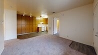 Located at Village Center Drive in Sacramento, CA, this 2-bedroom, 1-bathroom single-floor home offers a practical and comfortable layout. The open living area flows directly into the kitchen, creating a connected and functional space. The kitchen features warm oak-toned cabinetry, a full-size refrigerator, a dishwasher, a stove, and a stainless steel sink with light countertops, all set on wood-look vinyl flooring. A striking wall of floor-to-ceiling wood-panel cabinetry serves as a defining architectural feature between the living and kitchen areas, providing generous storage. The living area is finished with plush carpet and equipped with track lighting overhead. Both bedrooms are carpeted and bathed in natural light through large windows dressed with vertical blinds. The primary bedroom includes a dedicated closet area, while the second bedroom features sliding mirrored closet doors. The full bathroom is finished with wood-look vinyl flooring, a tub-shower combination with safety grab bars, and clean, neutral-toned walls. A utility room and additional closet space round out the home's practical offerings. The overall aesthetic is clean, neutral, and move-in ready.