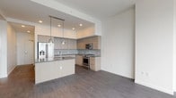 This contemporary single-floor residence showcases a clean, modern design with an open-concept layout that maximizes the sense of space. The kitchen is a standout feature, equipped with stainless steel appliances including a gas range, microwave, and French door refrigerator. Light wood cabinetry pairs elegantly with dark quartz countertops, while a central island with an undermount sink and pendant lighting above serves as both a functional workspace and a natural gathering point. Rich dark hardwood flooring extends throughout the main living and kitchen areas, adding warmth and contrast to the bright, white walls. The bedroom offers a serene retreat, carpeted for comfort and framed by large windows that frame expansive city skyline views, filling the room with natural light. A generously sized walk-in closet with built-in wire shelving provides ample storage. The bathroom is finished with neutral large-format floor tiles and subway-style wall tiles surrounding a full soaking tub and shower combination, complemented by a modern vanity with wood-toned cabinetry. Recessed lighting throughout the space enhances the clean, contemporary ambiance, making this a well-appointed and thoughtfully designed residence.