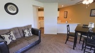This single-floor residence offers a practical and comfortable layout designed to maximize everyday living. The living area is carpeted and provides a warm, inviting atmosphere, accommodating casual seating and relaxed lounging. A partial dividing wall separates the living space from the kitchen zone while maintaining an open feel throughout.

The kitchen is equipped with white cabinetry, white appliances, and ample counter space, providing a clean and functional cooking environment. A breakfast bar with bar seating connects the kitchen to the living area, making it ideal for casual dining or entertaining. The accent wall in the kitchen area is painted in a warm earthy tone, adding a touch of character and color contrast to the otherwise neutral interior palette.

Recessed lighting in both the living and kitchen areas ensures consistent, even illumination throughout the space. The overall design is simple and clean, with white walls, neutral carpeting, and understated décor that creates a versatile backdrop adaptable to a variety of personal styles. The single-floor layout ensures easy navigation and accessibility throughout the entire residence.