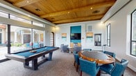 Located at 700 Cobia Dr in Katy, TX, this single-floor game and entertainment room is designed to deliver a refined yet relaxed recreational experience. The space is immediately distinguished by its warm, wood-paneled tray ceiling with recessed lighting, which adds both visual depth and a rich, inviting ambiance throughout.

At the heart of the room sits a full-size billiards table with a blue felt surface, complemented by a wall-mounted cue rack for convenient storage. A dedicated poker table surrounded by tufted blue leather chairs offers a sophisticated setting for card games and social gatherings. A large flat-screen television is mounted within a bold teal accent niche, accompanied by a media console and a compact under-counter refrigerator, making it ideal for entertainment viewing.

Comfortable lounge seating, including curved swivel chairs, is arranged throughout the space to encourage relaxed conversation and casual socializing. Tall arched windows line the walls, flooding the interior with generous natural light and framing views of the surrounding greenery. Large sliding glass doors open directly to an outdoor patio area, seamlessly connecting the indoor and outdoor spaces.

The neutral carpeted flooring ties the room together, while contemporary artwork adorns the walls, adding color and character. This versatile space is well-suited for community events, leisure, and social interaction.
