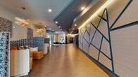 Located at 2865 NE Sandy Blvd in Portland, Oregon, this single-floor lobby space delivers a striking first impression through its thoughtfully curated contemporary design. The expansive corridor is anchored by warm wood-look flooring that runs the length of the space, complemented by a multi-level ceiling with recessed lighting and subtle LED accent strips that add depth and warmth throughout.

A standout architectural feature is the large geometric accent wall clad in angular, cream-toned panels outlined in dark teal, creating a bold visual focal point that sets the tone for the entire space. On the opposite side, generous windows invite natural light and offer views of the surrounding greenery, keeping the interior feeling open and connected to the outdoors.

The lounge area is furnished with vibrant mustard-yellow curved sectional seating, patterned upholstery, and sculptural wood pendant lights, creating an inviting and energetic gathering space. Curated wall art and layered textiles add personality and warmth to the seating nooks.

A dedicated refreshment station features clean white countertops, light wood cabinetry, stainless steel appliances, and a striking chevron-patterned teal tile backsplash. Elevator banks with brushed stainless steel doors and an Amazon Hub parcel locker round out the functional amenities, making this lobby a well-appointed and stylish hub for residents and visitors alike.