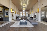 Located at 23400 Kingsland Blvd in Houston (Katy), TX, this upscale lobby and common area exudes modern sophistication across its generously sized single floor. The space is immediately defined by its dramatic double-height ceilings, which amplify the sense of openness and grandeur throughout. Tall, stone-clad columns rise from floor to ceiling, framing the open-plan layout and adding a refined architectural texture to the interior.

The flooring features a striking combination of large-format tile and rich wood-look planks, arranged in bold geometric patterns that guide the eye through the space. Ambient and accent lighting is a standout element, with oversized circular pendant fixtures suspended from a decoratively paneled ceiling trough, complemented by glowing amber wall sconces that cast a warm, inviting glow.

The central seating area showcases a large, contemporary sectional sofa in neutral grey tones, anchored by a multi-tiered rectangular coffee table atop a light area rug. Toward the rear, a staffed reception desk is set against a dramatically lit feature wall. A long communal table with modern chairs lines the window wall, bathed in natural light from floor-to-ceiling windows that offer views of the outdoors. Curated wall art and decorative accents complete the polished, welcoming atmosphere.