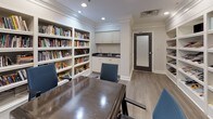 This single-floor space serves as a combined library and meeting room, thoughtfully designed to support both focused individual use and group collaboration. The room is finished with elegant crown molding, recessed lighting, and light wood-toned flooring that lends a clean, welcoming aesthetic throughout.

Floor-to-ceiling built-in white shelving lines the walls, generously stocked with an extensive collection of books and magazines, making the space a rich resource for reading and research. A large whiteboard mounted on the wall provides a practical focal point for presentations or brainstorming sessions, complemented by a dark wood conference table surrounded by ergonomic blue upholstered chairs that seat multiple participants comfortably.

A compact kitchenette is seamlessly integrated into the space, featuring white cabinetry with dark countertops and a sink — a convenient amenity for extended meetings or casual use. The room is accessed through a dark-framed frosted glass door, adding a touch of refinement while maintaining privacy. The overall ambiance balances functionality with a warm, library-like atmosphere, making it well-suited for reading, collaborative work, or small group meetings.