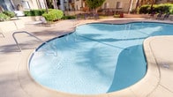 This outdoor amenity space offers a resort-style experience designed for relaxation and socializing. At its centerpiece is a generously sized freeform swimming pool with clear, blue-green water, accessible via stainless steel entry rails and featuring a gradual shallow end — ideal for a range of swimmers. Adjacent to the pool, a circular spa with active jets is nestled into a semi-enclosed alcove, surrounded by lush green hedging and furnished with rattan-style lounge chairs for post-soak relaxation.

The expansive sun deck is finished in light-toned concrete and provides ample open space for leisure. Multiple outdoor dining sets with umbrella-shaded tables and wrought iron chairs are arranged throughout the patio, offering comfortable spots to gather under the California sun. Mature palm trees and carefully maintained tropical landscaping frame the entire area, lending a lush, verdant atmosphere.

Dedicated BBQ stations equipped with full-size gas grills are positioned within gated alcoves along the perimeter, providing convenient outdoor cooking facilities. Ornamental iron fencing encloses the space, ensuring privacy while maintaining an open, airy feel. The surrounding multi-story buildings with tile rooflines give the complex a cohesive Mediterranean-inspired architectural character.