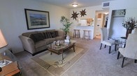 This single-floor residence offers a warm and inviting layout designed for comfortable everyday living. The living area is generously sized, featuring plush wall-to-wall carpeting, a large comfortable sofa, and a glass-topped coffee table set atop a decorative area rug. A framed landscape artwork anchors one wall, while decorative star accents add character above the open passage to the kitchen. Natural and ambient lighting keep the space feeling bright and welcoming throughout.

The kitchen is connected to the living area via an open breakfast bar, which is lined with wooden barstools and provides a convenient casual dining option. The kitchen itself is equipped with standard appliances, including a refrigerator and range, and features warm wood-toned cabinetry that adds a cozy feel. The open-concept design between the living room and kitchen encourages an easy flow for both entertaining and daily routines.

The overall ambiance of the space is relaxed and functional, with neutral tones in the walls, flooring, and furnishings that create a cohesive and understated aesthetic. The single-floor layout ensures easy accessibility throughout, making this a practical and comfortable home.