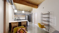 Located at 300 N Cherapa Place in Sioux Falls, SD, this single-floor unit offers a thoughtfully designed loft-style living experience. The open-concept main living area immediately impresses with its warm wood-planked ceiling, exposed industrial ductwork, and polished concrete floors — a striking combination that balances raw character with modern refinement. Pendant globe lighting adds a soft, inviting glow throughout.

The space features two distinct kitchen areas, both outfitted with gray cabinetry, white countertops, stainless steel appliances including full-size refrigerators, and open wood shelving for display and storage. A kitchen island with woven bar seating anchors the main kitchen, making it ideal for casual dining and entertaining.

A dedicated workspace with a lean shelving unit and seating is neatly integrated into the living area, maximizing the functionality of the layout. The spacious primary bedroom opens to an outdoor balcony, filling the room with natural light, and includes an in-unit washer conveniently tucked behind a sliding barn-style door.

The bathroom features clean, modern finishes including hexagonal white floor tile, a dark-toned vanity with white countertop, and warm neutral walls. Every detail of this unit reflects a cohesive industrial-modern aesthetic.
