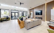 Located in Miami, FL, this sleek, single-floor communal space delivers a sophisticated open-plan layout that seamlessly integrates a fully equipped kitchen and an expansive living area. The interior is defined by soaring ceilings with clerestory windows that flood the space with abundant natural light, complemented by recessed lighting and modern ceiling fans. Large floor-to-ceiling glass doors and windows frame views of the outdoor surroundings, enhancing the connection between interior and exterior.

The kitchen features a generous white quartz island with seating for several bar-stool guests, warm wood-paneled cabinetry with open shelving, stainless steel appliances, and a sleek linear pendant light overhead. Adjacent to the kitchen, a casual lounge area offers an olive-green sectional sofa and round bistro tables, ideal for informal gatherings.

The living room is anchored by a striking floor-to-ceiling slatted wood accent wall housing a wall-mounted television above a linear fireplace. Comfortable neutral seating arrangements, accent chairs in complementary tones, and curated artwork contribute to the refined yet welcoming ambiance. Polished large-format marble-look tile flooring flows throughout, tying the entire space together with an air of modern elegance.