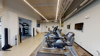 Located at 1301 E Washington St in Indianapolis, Indiana, this fitness center occupies a single-floor space with a distinct industrial character. The facility's architecture retains exposed structural trusses, open ductwork, and polished terrazzo flooring throughout the main areas, giving it a converted warehouse aesthetic. Linear LED lighting runs along the ceiling, providing bright, even illumination across the entire space.

The cardio zone is a central feature, housing a row of Matrix treadmills, elliptical trainers, and a rowing machine, all positioned on a dedicated flooring platform. Wall-mounted flat-screen televisions are positioned for clear viewing during workouts, and a water fountain is accessible nearby.

A dedicated functional training alcove offers a heavy punching bag, battle ropes, a medicine ball, and a well-stocked weight rack with kettlebells and additional equipment. Large steel-framed windows in this area allow natural light to enter and provide outdoor views of the surrounding greenery.

A separate room houses stationary cycling bikes, also benefiting from generous natural light through floor-to-ceiling industrial-style windows. The entry area features terrazzo floors, locker storage, and a towel station, providing a practical transition into the workout space. Overall, the facility presents a functional, no-frills environment suited for comprehensive fitness training.