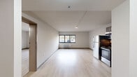 This single-floor unit features a clean, modern design across a thoughtfully arranged layout that includes an open-concept kitchen and living area, a separate living room, two bathrooms, and a closet. Light-toned luxury vinyl plank flooring runs throughout the main living spaces, lending a bright, cohesive feel to the interior. The open kitchen is equipped with stainless steel appliances — including a refrigerator, range, and microwave — paired with dark wood cabinetry and a simple countertop with an integrated sink, offering a functional and contemporary cooking space. The living area is airy and open, with neutral-toned walls and a large window that allows natural light to fill the room while providing an outdoor view. A wall-mounted HVAC unit ensures year-round comfort. The separate living room provides additional flexible living space with the same light flooring and clean wall finishes. The two bathrooms are finished in warm-toned ceramic tile featuring a decorative diamond accent border, with granite countertops, undermount sinks, and chrome fixtures. A bathtub with a curved shower rod is present in one bathroom. The overall ambiance is modern, functional, and move-in ready, with a neutral palette that provides a versatile backdrop for a variety of interior styles.