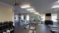 This fitness room is designed to support a comprehensive workout routine within a single-level space. The room features durable dark rubber flooring throughout, providing a practical and resilient surface suited for exercise. Overhead, fluorescent lighting fixtures ensure the space is well-lit, complemented by ceiling fans that promote air circulation during workouts. Multiple windows fitted with white blinds line the walls, allowing natural light to filter into the room while maintaining privacy.

The space is equipped with a range of cardio machines, including a stationary bike and an elliptical trainer, ideal for cardiovascular conditioning. A multi-function cable machine serves as the centerpiece for strength training, surrounded by free weights and adjustable benches that accommodate a variety of exercises. A weight plate storage rack and additional free weights are neatly organized along one wall.

A large floor-to-ceiling mirror spans a portion of one wall, expanding the sense of space and allowing users to monitor their form. Crown molding adds a refined architectural touch. The overall layout is open and functional, offering ample room to move between equipment comfortably.