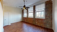 This one-bedroom, one-bathroom unit in New Orleans blends historic architectural character with functional modern living. The main living area is defined by a striking exposed brick wall punctuated by multiple windows that frame views of the downtown New Orleans skyline, flooding the space with natural light. Rich hardwood flooring runs throughout, complemented by a white wood-paneled ceiling and a ceiling fan overhead, creating a warm, loft-inspired atmosphere.

A dedicated hallway connects the living area to the remaining rooms, featuring a louvered closet door for storage and staircase access. The kitchen is efficiently laid out with warm wood cabinetry, black appliances including a range, microwave, and refrigerator, and hardwood flooring that carries through from the main living space. Pendant lighting adds a contemporary touch above.

The bathroom includes a full tub-shower combination with clean white tile surround. A dedicated in-unit laundry closet houses a stacked washer and dryer, offering added convenience. An additional open room with a ceiling fan and hardwood floors provides flexible use as a bedroom or secondary living space, complete with a decorative column detail. Overall, the unit delivers a balance of historic New Orleans charm and practical urban living.