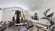 Located at 2010B Pinehurst Court in Allentown, PA, this dedicated fitness center offers a well-equipped exercise space designed to support a full range of workout routines. The room features a vaulted ceiling that enhances the sense of openness, complemented by recessed lighting and several horizontal windows that invite natural light from the surrounding greenery. The interior is finished with clean white walls, a combination of rubber mat and carpet flooring, and a large wall-mounted mirror that visually expands the space and aids in form monitoring during exercise. The cardio zone is outfitted with a treadmill, elliptical trainer, and stationary bike — all Spirit-branded — alongside a wall-mounted flat-screen television for entertainment during workouts. The strength training area includes a multi-station cable machine, adjustable benches, and a tiered dumbbell rack with a comprehensive range of free weights. A wall-mounted split air conditioning unit ensures year-round climate control, while a baseboard heater provides supplemental warmth. Additional amenities include a paper towel dispenser, waste bin, and a keyed entry door with an exterior window panel. The space is compact yet thoughtfully organized to maximize functionality for residents seeking convenient, on-site fitness access.