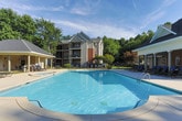 This single-story amenity space in Newnan, Georgia, is designed to support an active lifestyle through two distinct areas: a comprehensive fitness center and an outdoor aquatic area. The fitness center is well-lit by large, multi-pane windows that invite abundant natural light and offer views of the surrounding greenery. Full-length mirrors line portions of the walls, visually expanding the space while allowing users to monitor their form. The cardio area is equipped with treadmills, elliptical machines, and a stationary spin bike, complemented by wall-mounted televisions for entertainment during workouts. A separate section houses a Concept2 rowing machine and a cable weight machine, providing full-body strength training options. An additional free-weight area features a tiered dumbbell rack, kettlebells, medicine balls, and a adjustable workout bench, catering to varied training needs. Restroom facilities are conveniently accessible within the fitness center. The outdoor pool area features a generously sized, curved swimming pool with clear blue water, surrounded by a wide concrete deck lined with lounge chairs. A white pergola provides a shaded retreat, while mature trees create a natural, private backdrop.