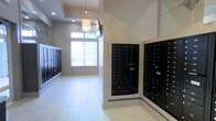 This single-floor space serves as a functional and polished mail center and entry area, designed to accommodate a large number of residents with ease and efficiency. The room is lined on multiple walls with built-in black metal mailbox units, neatly organized in rows and columns to provide ample individual storage for correspondence and packages. The layout is thoughtfully arranged to allow comfortable circulation throughout the space.

The interior features light-toned tile flooring that reflects natural light, contributing to a clean and open atmosphere. Walls are finished in a warm neutral tone, complementing the crisp white baseboards and architectural column details. A large window with horizontal blinds allows natural light to filter into the space, enhancing the bright and welcoming ambiance.

Overhead, recessed lighting is complemented by a decorative pendant chandelier, adding a touch of refinement to what is otherwise a utilitarian space. The raised ceiling with a tray detail further elevates the room's sense of height and openness. A wall-mounted monitor is also present, suggesting the space may serve an informational or security function as well. Overall, the area balances practicality with a polished aesthetic, creating a tidy and inviting environment for daily resident use.