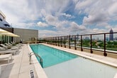 Perched high above Atlanta's Old Fourth Ward neighborhood, this rooftop amenity deck at 639 Glen Iris Dr NE offers a striking combination of resort-style relaxation and urban panoramas. The centerpiece is a generously sized lap pool finished in light tile, flanked on both sides by a wide sun deck lined with contemporary white lounge chairs and market umbrellas. A glass-panel railing runs the perimeter of the rooftop, preserving unobstructed, sweeping views of the Atlanta skyline to one side and lush treetop canopy to the other. Floor-to-ceiling bronze-framed glass walls enclose an interior lounge area adjacent to the pool deck, blurring the boundary between indoor and outdoor space and allowing natural light to flood both areas. A dedicated alfresco seating nook features a round table with wicker chairs, surrounded by ornamental grasses and planters, creating an intimate outdoor dining or gathering spot. The deck's light-toned porcelain tile flooring ties the entire space together with a clean, modern aesthetic. Together, the pool, lounge areas, and panoramic city views make this rooftop deck a premier outdoor amenity in the heart of Atlanta.