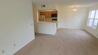 This single-floor residence features an open-concept design that seamlessly connects the living area to the kitchen, creating a functional and cohesive layout. The living area is generously sized, finished with wall-to-wall neutral-toned carpeting that lends a clean, understated look throughout the space. White walls and white baseboards contribute to a bright, airy atmosphere.

The kitchen is positioned centrally and elevated slightly behind a raised peninsula counter, which serves as both a workspace and a casual dining or serving area. Warm-toned wooden cabinetry lines the kitchen walls, complemented by a white refrigerator, a black microwave, and a dark cooktop. The peninsula provides convenient pass-through access, making the kitchen feel connected to the main living space without sacrificing definition.

Overhead, a modest chandelier with multiple candelabra-style bulbs adds a decorative touch to the living area. Ventilation is provided by a ceiling vent visible above the kitchen. The overall palette — neutral carpet, white walls, and warm wood cabinetry — gives the space a simple, move-in-ready character suited to a variety of personal styles.