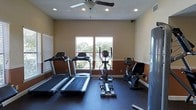 This single-level fitness room offers a dedicated workout space equipped with a thoughtful selection of cardio machines. The room features two treadmills positioned toward the front, an elliptical trainer centrally placed, and a stationary recumbent-style bike along the right side, providing a variety of cardiovascular exercise options. The flooring is finished with dark rubber matting, which is both durable and practical for a fitness environment. Natural light enters through two windows along the back wall, offering views of surrounding greenery and contributing to an open, airy feel. A ceiling fan mounted overhead ensures comfortable air circulation during workouts. The walls feature a two-tone paint scheme, with a warm tan upper half and a deeper reddish-brown lower half, divided by a subtle chair rail, adding a degree of visual interest to the otherwise functional space. Recessed lighting supplements the natural light, ensuring the room remains well-lit at all times. White baseboards and a clean, minimal layout complete the room's straightforward, utilitarian aesthetic, making it a practical and well-appointed space for regular fitness use.