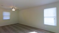 This single-floor space presents a straightforward and functional layout designed for comfortable everyday living. The main living area is finished with wall-to-wall carpeting that adds warmth underfoot and contributes to a quiet, cozy atmosphere. Neutral-toned walls keep the space feeling bright and open, providing a versatile backdrop for a variety of interior styles and furnishings. A ceiling fan with an integrated light fixture is centrally mounted, offering both overhead illumination and air circulation throughout the room. Natural light filters in through a window fitted with horizontal blinds, allowing for easy adjustment of light and privacy as needed. Clean white baseboards frame the perimeter of the room, adding a tidy, finished look to the overall space. The open floor plan allows for flexible furniture arrangement, making the area adaptable to different uses. The overall ambiance is clean, neutral, and move-in ready, suited for those seeking a simple and practical living environment.