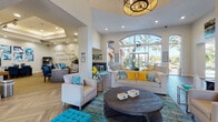 Located at 2450 Lake Debra Dr in Orlando, FL, this single-story leasing office and community clubhouse exudes a warm, coastal-inspired atmosphere throughout its thoughtfully designed interior. The grand main lobby draws immediate attention with its soaring ceilings, dramatic arched floor-to-ceiling windows, and an abundance of natural light that frames views of the surrounding palm-lined exterior. Rich herringbone-patterned hardwood flooring runs consistently throughout, anchoring the space with a refined yet inviting aesthetic.

The main lounge is furnished with cream upholstered seating, teal accent ottomans, and bold yellow throw pillows, complemented by a substantial round wooden coffee table and a layered teal area rug. Statement chandelier lighting overhead adds an elegant touch. An adjacent leasing office area features workstations and is accented with inspirational wall typography, reinforcing a resident-first philosophy.

A secondary lounge or waiting room offers a more intimate setting with wall-mounted TV, yellow curtains, herringbone floors, and a water station for guest comfort. Frosted glass sliding panels create flexible, semi-private spaces that maintain an open, airy feel. A dedicated children's activity nook includes a small table set, bean bag seating, and a pass-through window overlooking the main lobby, making the facility welcoming for families.