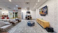 Located at 3333 Wisconsin Ave NW in Washington, DC, this thoughtfully designed community lounge offers a generous, open floor plan that effortlessly accommodates a variety of activities and social configurations. The space is anchored by a large communal wood-top table with sleek pendant lighting overhead, providing an ideal setting for collaborative work or group gatherings. Warm wood-clad columns punctuate the open layout, adding architectural interest and naturally defining distinct zones throughout the room.

A spacious lounge area features a large sectional sofa accented with vibrant throw pillows, complemented by stylish teal accent chairs arranged near a striking decorative screen partition. A dedicated reading nook, lined with colorful bookshelves and comfortable lounge chairs, invites quiet relaxation. The media zone is equipped with computer workstations, wall-mounted displays, and a tufted leather ottoman, offering both productivity and entertainment options.

Exposed brick-effect wall panels and light wood finishes lend a modern yet welcoming ambiance throughout. Soft, patterned carpeting ties the space together underfoot, while recessed lighting provides consistent, comfortable illumination. A kitchenette area is visible in the background, rounding out this well-appointed communal space with everyday convenience.