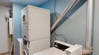 This single-floor utility space serves a dual purpose, functioning as both a laundry room and a utility/mechanical room. The laundry area is equipped with commercial-grade Speed Queen top-loading washers and stacked dryer units, offering efficient, heavy-duty washing and drying capacity. A deep utility sink with exposed plumbing and shut-off valves provides additional functionality, while a large ventilation duct runs diagonally along the wall, highlighting the room's industrial character. The walls in the laundry section are painted in a light blue tone, giving the area a distinct visual identity.

The utility section transitions into a clean, white-walled space featuring a wall-mounted built-in countertop — ideal for folding or workspace use — alongside an electrical outlet. Dominating one full wall of the room is an extensive bank of individual electrical meters, serving what appears to be a multi-unit building. The meters are mounted in large gray panels and represent the central electrical management hub for the property. The floor throughout is finished with resilient tile. Overhead flush-mount lighting provides consistent illumination. Together, the two areas form a functional, no-frills utility core built for practical, day-to-day operational needs.