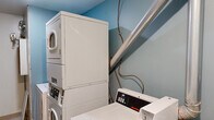 This single-floor utility space serves a dual purpose, functioning as both a laundry room and a utility/mechanical room. The laundry area is equipped with commercial-grade Speed Queen top-loading washers and stacked dryer units, offering efficient, heavy-duty washing and drying capacity. A deep utility sink with exposed plumbing and shut-off valves provides additional functionality, while a large ventilation duct runs diagonally along the wall, highlighting the room's industrial character. The walls in the laundry section are painted in a light blue tone, giving the area a distinct visual identity.

The utility section transitions into a clean, white-walled space featuring a wall-mounted built-in countertop — ideal for folding or workspace use — alongside an electrical outlet. Dominating one full wall of the room is an extensive bank of individual electrical meters, serving what appears to be a multi-unit building. The meters are mounted in large gray panels and represent the central electrical management hub for the property. The floor throughout is finished with resilient tile. Overhead flush-mount lighting provides consistent illumination. Together, the two areas form a functional, no-frills utility core built for practical, day-to-day operational needs.