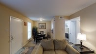This single-floor residence presents an open-concept layout that seamlessly connects the living, dining, and kitchen areas into one cohesive, functional space. The living area is anchored by a comfortable sofa positioned on a patterned area rug, providing a welcoming focal point for the room. Warm beige walls and light-toned ceilings create a bright, neutral atmosphere throughout the space.

Adjacent to the living area, the dining space accommodates a dark-wood table and chair set, positioned near a window that allows natural light to filter into the room. A framed artwork piece adorns the wall above, adding a decorative touch to the dining area. A tall indoor plant nearby adds a natural, organic element to the space.

A partial wall with a countertop pass-through connects the dining area to the kitchen, which is visible from the main living space. The kitchen features white cabinetry and appliances, including a stove and microwave, offering a clean, practical cooking environment. Overhead ventilation units are present throughout, ensuring comfortable climate control. The overall ambiance is casual, practical, and well-organized.