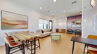 Located at 5900 N 99th Ave in Glendale, Arizona, this single-floor communal space offers a well-designed open-plan layout that seamlessly blends a living area with a dining zone. Light-toned hardwood-style flooring runs throughout, complementing the crisp white walls and lending a clean, modern aesthetic to the entire space.

The living area is anchored by a tufted beige sofa adorned with colorful accent pillows, positioned beneath a window fitted with a roller shade that diffuses natural light. A round marble coffee table and a sculptural green lounge chair add character, while curated artwork enhances the walls. Elegant pendant lighting descends from the ceiling in the transitional zone, highlighting a warm-toned wood credenza and a soft lavender accent wall that adds a touch of color.

The dining area features multiple seating configurations, including a striped high-top communal table with bar-height chairs and smaller bistro-style tables paired with mustard-yellow modern chairs. Large black-framed windows frame views of the surrounding courtyard, flooding the space with abundant natural light. Stylish cylindrical glass pendant lights hang above the dining tables, adding a refined focal point.

Overall, the space strikes a balance between functionality and contemporary style, making it an inviting and versatile environment for both casual gatherings and focused activities.