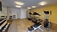 This fitness center is a thoughtfully designed, single-floor facility offering a comprehensive workout environment. The space is finished with neutral warm-toned walls and a combination of tile and carpet flooring, creating a clean and welcoming atmosphere throughout.

The main cardio area is well-lit by multiple flush-mount ceiling fixtures and features multiple treadmills, an elliptical trainer, a stationary bike, and a recumbent bike, providing a full range of cardio options. A wall-mounted split air conditioning unit ensures a comfortable climate during workouts. Large sliding glass doors along one wall flood the space with natural light and open to an outdoor patio area, offering a pleasant connection to the exterior.

A dedicated free weights section includes a full rack of dumbbells and is enhanced by a large wall mirror that visually expands the space. A wall-mounted television adds entertainment options during workouts. An adjustable workout bench serves both the cardio and free weights zones. A standing physician's scale is conveniently positioned near the entry area for tracking fitness progress.

The facility also includes a separate door leading to an additional room, providing functional utility space. Exercise reference posters and seating round out the amenities, making this fitness center a complete and practical space for a wide range of exercise routines.