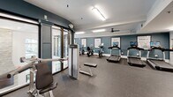 Located at 7000 Johnson Farm Ln in Chadds Ford, PA, this fitness center is a thoughtfully designed, single-floor facility that offers a comprehensive workout experience. The space is defined by its clean, modern aesthetic, featuring teal accent walls, crisp white crown molding, and durable rubber flooring throughout. Motivational framed typography — with phrases like "Spin," "Run," "Rest," and "Crunch" — adorns the walls, reinforcing the energetic atmosphere.

The cardio zone is generously equipped with multiple treadmills and stationary bikes, each outfitted with individual screens for an engaging workout experience. A dedicated cycling and elliptical area provides additional low-impact training options, complemented by natural light from large windows overlooking lush greenery. Ceiling fans contribute to a comfortable environment during intense sessions.

The strength training area features a full rack of dumbbells, weight benches, and a multi-station cable machine, all set against a floor-to-ceiling mirror wall that amplifies the sense of space and allows for proper form checks. Recessed and overhead fluorescent lighting ensures the entire facility is well-illuminated. A seating bench and utility station near the exit add functional convenience, making this fitness center a complete and inviting space for all levels of exercise.