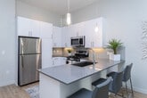Located at 132 E Midvillage Blvd in Sandy, UT, this well-designed one-bedroom, one-bathroom unit offers a thoughtfully organized single-floor layout. The open-concept kitchen and living area creates a seamless flow, anchored by a sleek kitchen featuring white cabinetry, a subway tile backsplash, stainless steel appliances including a gas range, built-in microwave, refrigerator, and dishwasher, along with a kitchen island with pendant lighting overhead. Light wood flooring runs throughout the main living areas, adding warmth and continuity. Large windows dressed with sheer white curtains allow generous natural light to fill the living room, which benefits from elevated ceilings that enhance the sense of openness. The entry hallway provides a welcoming transition into the main living space. The bedroom features plush carpet flooring, a bright window with blinds, and direct access to a spacious walk-in closet outfitted with multi-level wire shelving and ample hanging space. The bathroom is finished with wood-toned cabinetry, a white quartz countertop, a large mirror, and a full tub-shower combination with white subway tile surround and light wood-style flooring. Every room reflects a clean, modern aesthetic throughout.