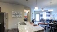 This single-floor residence showcases a thoughtfully designed open-concept layout that connects the kitchen, living area, and bedroom with ease. The kitchen is anchored by a generously sized granite-topped island with a double undermount sink, offering both functional workspace and casual dining space. Elegant pendant lights with bell-shaped shades hang above the island, providing warm, focused illumination. The neutral gray walls create a cohesive backdrop throughout the space, complementing the wood-style flooring that runs underfoot.

The living area extends naturally from the kitchen, benefiting from natural light streaming through a glass door that leads to an outdoor space. A ceiling fan with an integrated light fixture ensures comfortable airflow. Striped navy and white curtains frame the door, adding a decorative touch. A glimpse into the adjacent bedroom reveals a cozy, well-appointed space with a dark-toned bed frame and warm accent colors. The open sightlines between rooms enhance the sense of spaciousness, making the layout feel connected and functional throughout.