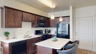This single-floor residential unit offers a thoughtfully designed layout that balances functionality and comfort across its key living spaces. The kitchen serves as a central highlight, featuring rich dark wood cabinetry with brushed nickel hardware, white countertops, and a full suite of black appliances including a gas range, built-in microwave, dishwasher, and refrigerator. A peninsula with seating provides a casual dining option and open sightlines into the living area. Pendant lighting above the peninsula adds a modern touch, while wood-look flooring grounds the space with warmth. The living room benefits from multiple windows that invite generous natural light and is complemented by a ceiling fan. The two bedrooms offer a quiet retreat, each featuring carpet flooring, neutral walls, and windows with blinds that allow for controlled light. The full bathroom is finished with a dark wood vanity, light countertops, wood-look flooring, and a tub-shower combination. Throughout the unit, a consistent design palette of warm neutrals and dark wood tones creates a cohesive and inviting atmosphere suited for everyday living.
