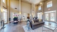 This single-floor community clubhouse or leasing center is defined by its dramatic double-height ceilings, which create an airy, grand ambiance throughout the main gathering area. Warm taupe and gray tones coat the walls, complemented by wide-plank wood-style flooring that runs seamlessly across the space. The main lounge area features a plush gray sofa, area rug, and accent tables beneath a striking geometric orb chandelier, establishing an inviting focal point for residents and visitors alike.

Adjacent to the lounge, a dining area hosts a sleek glass-top table with black chairs and vibrant orchid arrangements, adding a pop of color against the neutral palette. Tall French doors and transom windows line the perimeter, flooding the interior with abundant natural light while offering views to the outdoors.

A cozy sitting nook is anchored by a fireplace with rich wood built-ins displaying decorative accents, a wall-mounted television, and a patterned accent chair. Nearby, a dedicated workspace features a wood writing desk with chair, ideal for focused tasks. A stylish beverage station with a coffee maker provides a welcoming amenity. Private office spaces enclosed by glass-paned French doors are visible along the perimeter, supporting administrative or leasing functions. The overall atmosphere is polished, functional, and warmly welcoming.